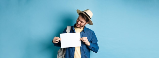 Free photo happy tourist in straw hat smiling and looking at piece of paper for your logo standing on blue back