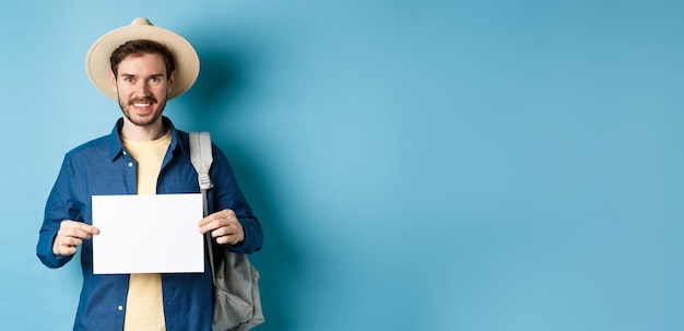 Free photo happy tourist with backpack showing empty piece of paper smiling at camera standing on blue backgrou