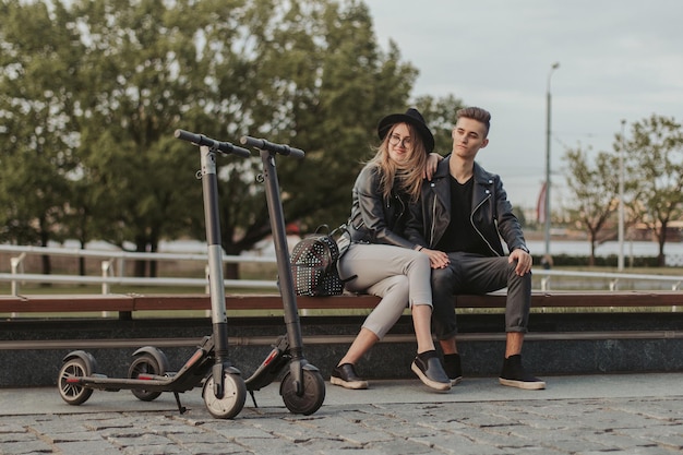 Happy trendy couple is chilling on the bench at city park with their scooters.