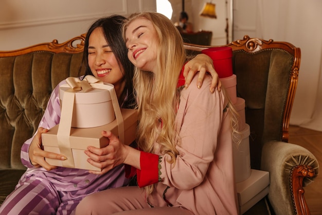 Happy two interracial young girls are sitting hugging on couch and holding gift boxes Emotion concept of people at birthday party