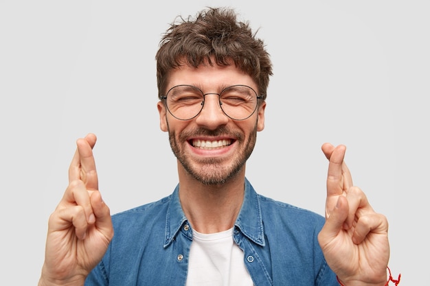 Free photo happy unshaven male with broad smile, shows white teeth, crosses fingers for good luck, being in high spirit stands over white wall wears fashionable denim shirt. positive guy makes hope gesture