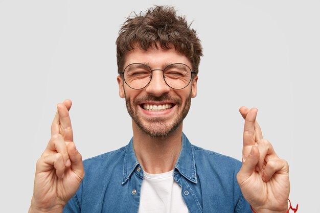 Happy unshaven male with broad smile, shows white teeth, crosses fingers for good luck, being in high spirit stands over white wall wears fashionable denim shirt. Positive guy makes hope gesture