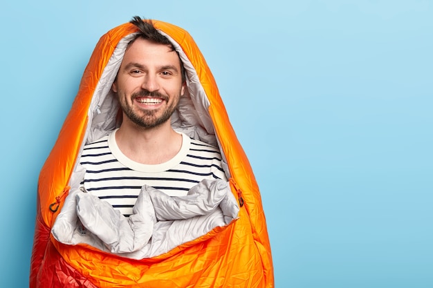 Free Photo happy unshaven man vacationist spends holidays near mountains in camping, sleeps in sleeping bag, smiles positively, dressed in striped sweater, poses against blue wall