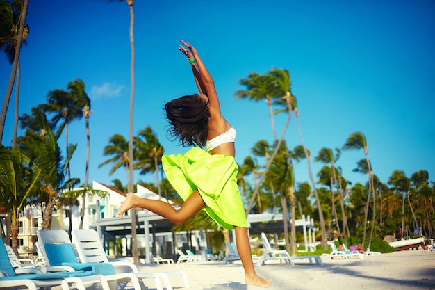 Free Photo happy urban modern young stylish woman woman model in bright modern cloth in green colorful skirt outdoors in the summer beach jumping behind blue sky