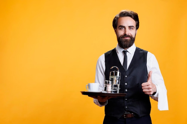 Free photo happy waiter does thumbs up sign while he holds platter with coffee and accessories in studio, professional stylish butler. man restaurant employee showing like agreement gesture on camera.