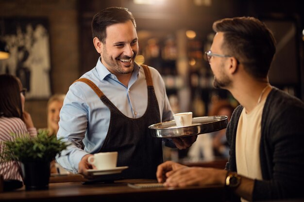 Happy waiter serving coffee and communicating with male guest in a bar