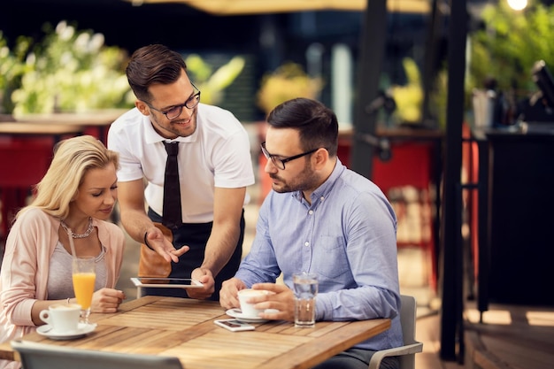 Happy waiter talking to guests while using digital tablet and showing them the menu in a restaurant