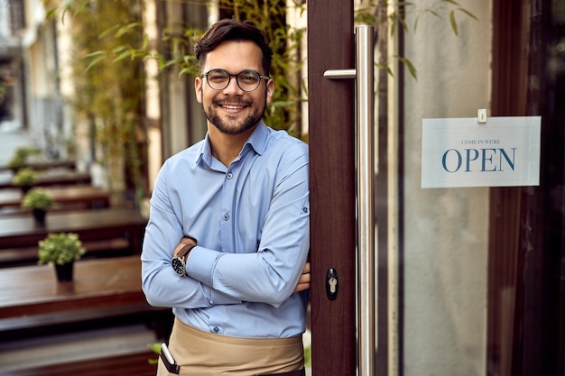 Free photo happy waiter with arms crossed standing next to open sign at the cafe's entrance and looking at the camera