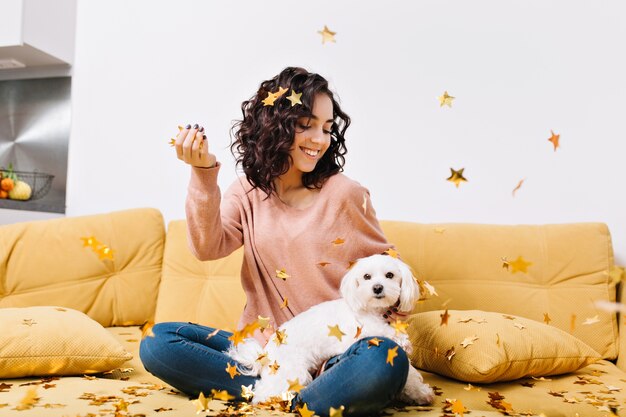 Happy weekends, true positive emotions of young joyful woman with cut curly hair having fun with little dog in falling golden tinsels on couch in modern apartment