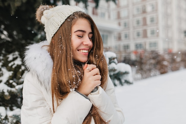 Happy winter time of young joyful woman enjoying snow in city. Attractive woman, long brunette hair, smiling with closed eyes.