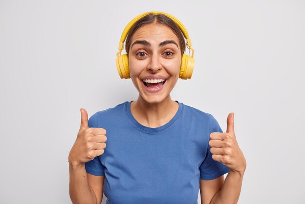 Happy woman approves something keeps thumbs up recommends to listen this audio track wears stereo yellow headphones on ears dressed in blue t shirt isolated over white background. I like it.