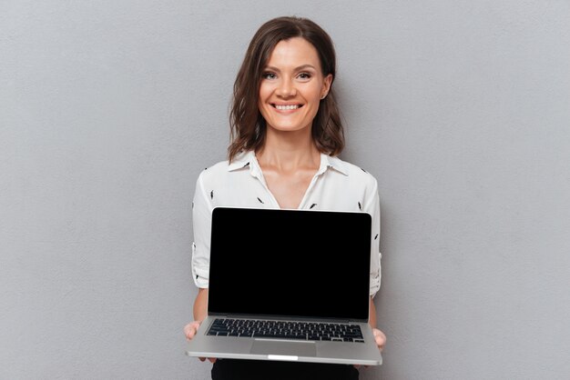 Happy woman in business clothes showing blank laptop computer screen on gray