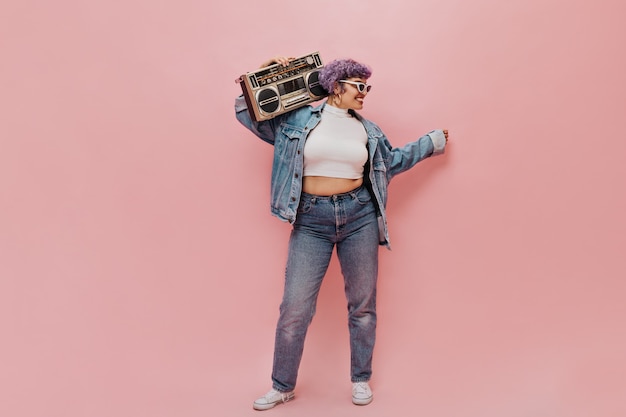 Free photo happy woman in denim wide jacket and tight pants posing on pink. stylish woman in white sunglasses holds tape recorder.