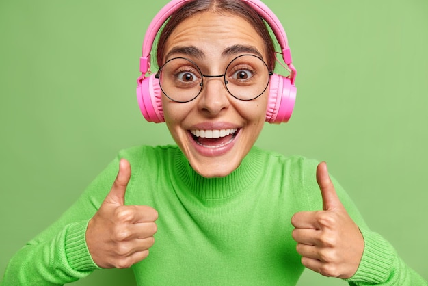 Free Photo happy woman enjoys nice audio track does positive gesture with hands keeps thumbs up says i like it wears audio track in pink headphones wears casual clothes round spectaclesisolated on green wall