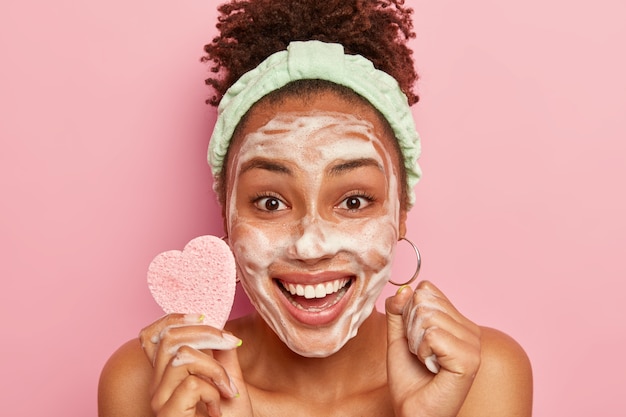 Free Photo happy woman enjoys relaxing time, washes face with soap bubble, feels refreshed and delighted, holds cosmetic sponge for wiping complexion