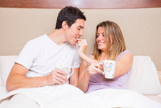 Happy woman feeding cookie to her boyfriend sitting on bed