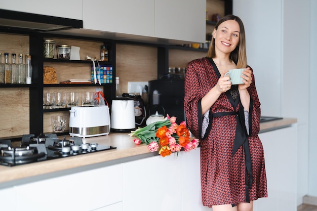 Free photo happy woman having a coffee at home in the kitchen wearing a silk robe while enjoying fresh flowers