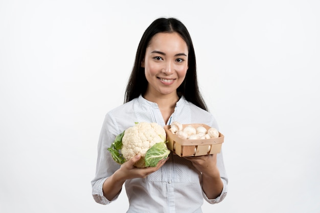 Happy woman holding raw fresh cauliflower and mushrooms in front of white backdrop