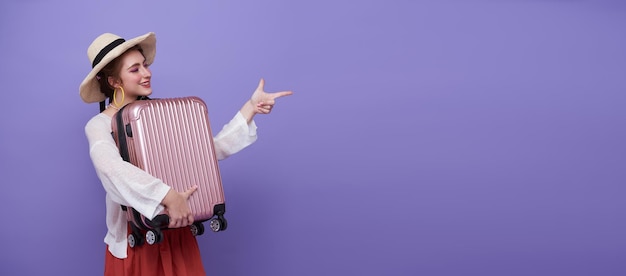 Happy woman holding suitcase and pointing her finger on purple background