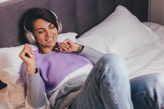 Happy woman at home on comfy bed wearing warm clothes pullover, listen music