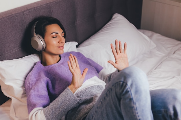 Happy woman at home on comfy bed wearing warm clothes pullover, listen music