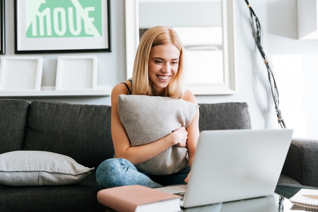 Happy woman hugging pillow and using laptop on sofa