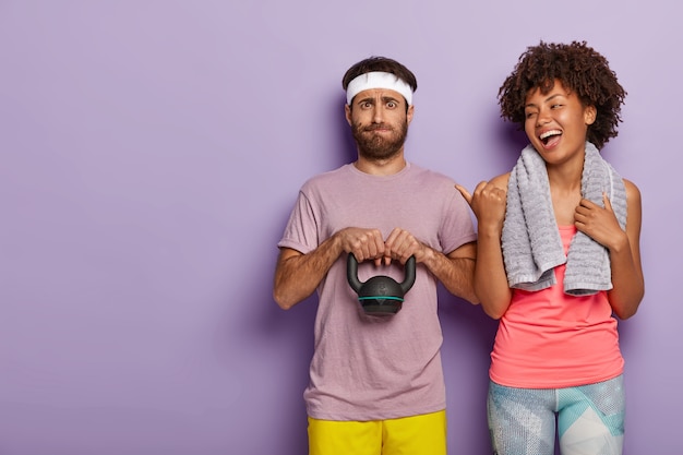 Free photo happy woman laughs sincerely, points away with thumb, indicates at boyfriend who lifts weight