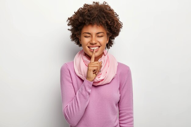 Happy woman makes silence gesture, keeps index finger over lips, glad to share pleasant secret with best friend, wears purple casual jumper