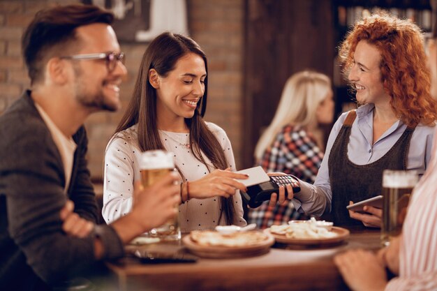 Happy woman paying to a waitress via contactless smart phone payment in a bar