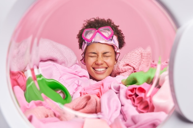 Free photo happy woman puts laundry in washing machine does housework smiles broadly drowned in heap of unwashed clothes