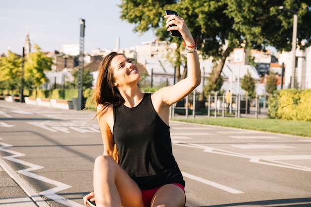 Happy woman sitting on skateboard taking selfie with cellphone