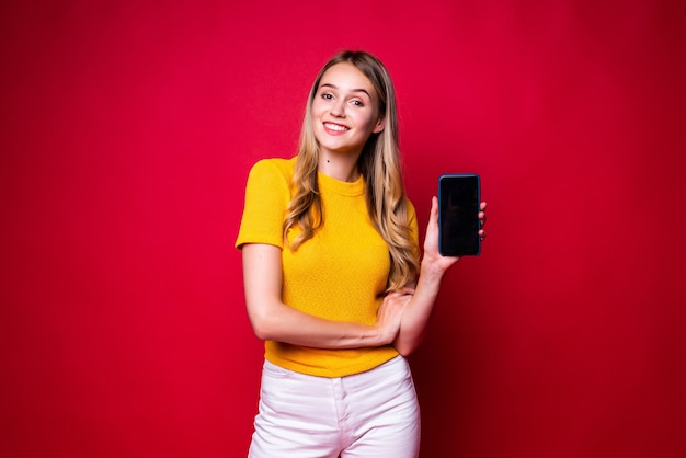 Happy woman smiling and gesturing finger aside on black screen of cell phone isolated over red wall