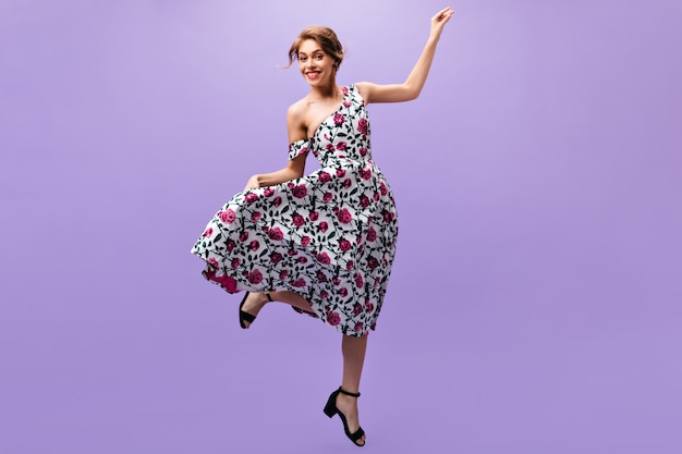 Happy woman in stylish dress jumps on purple background. Joyful attractive girl in floral trendy outfit posing on isolated backdrop.