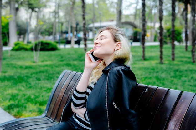 Happy woman talking on mobile phone wearing black leather jacket and sitting outdoor on a bench