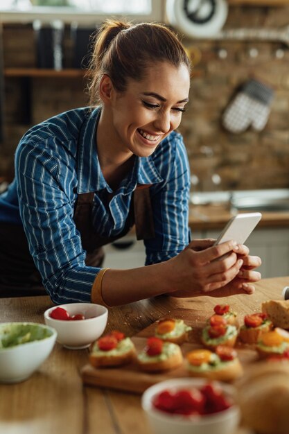 Happy woman using smart phone and photographing food she has prepared in the kitchen