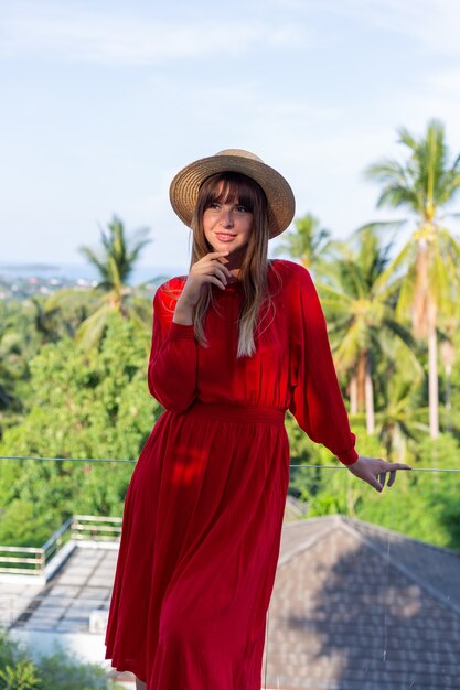 Happy woman on vacation in red summer dress and straw hat on balcony with tropical view on sea and plam trees.