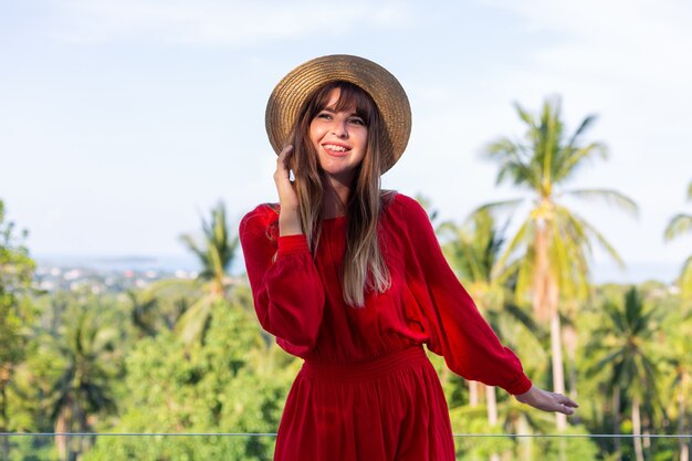 Happy woman on vacation in red summer dress and straw hat on balcony with tropical view on sea and plam trees.