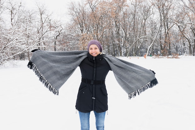 Happy woman wearing warm clothes and holding cozy scarf standing on snowy land in winter