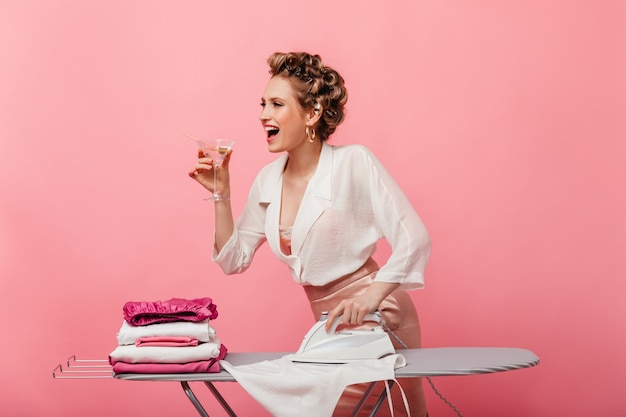 Happy woman in white blouse and pink skirt holding martini glass and iron