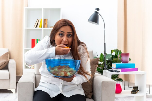 Happy woman in white shirt and black pants relaxing sitting on the chair with bowl of chips eating chips in light living room