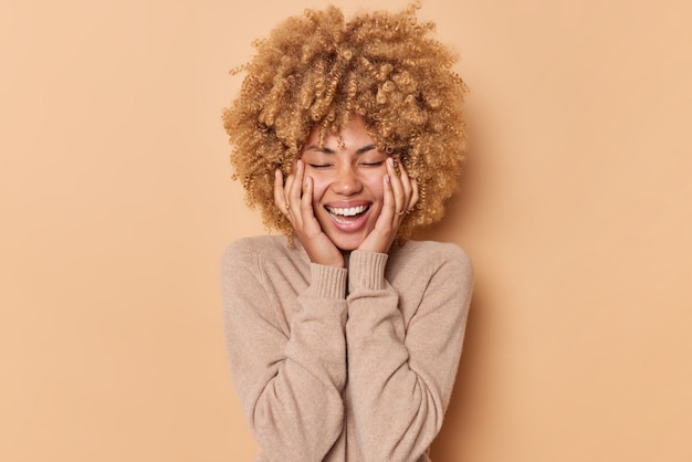 Free photo happy woman with curly hair keeps hands on cheeks closes eyes and smiles joyfully wears casual jumper being in good mood isolated over beige background recalls nice memories. emotions concept