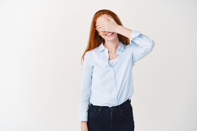 Happy woman with ginger hair covering eyes with hand and smiling carefree, waiting for surprise, standing over white wall