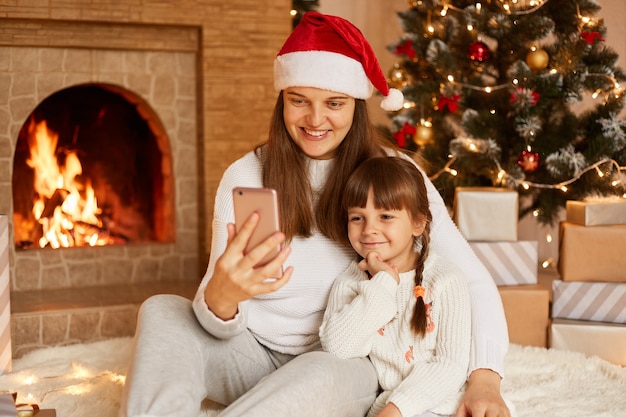 Happy woman with her cute little daughter sitting on floor near Christmas tree and fireplace, holding smart phone, looking at device screen, having positive facial expressions and festive mood.