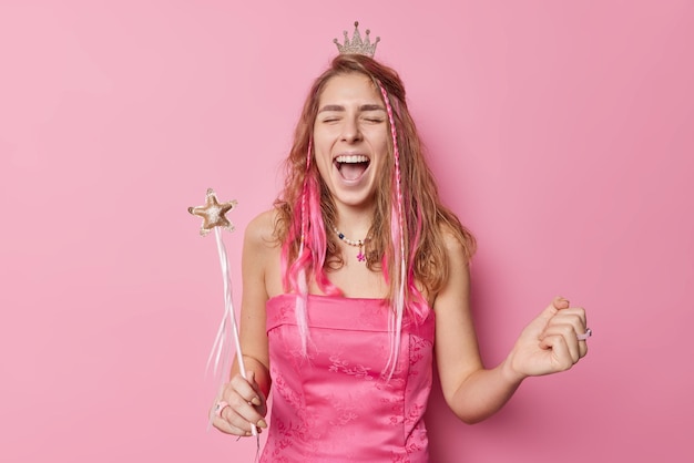 Happy woman with long hair exclaims loudly clenches fist keeps mouth widely opened holds magic wand wears dress and crown celebrates something isolated over pink background Holiday concept