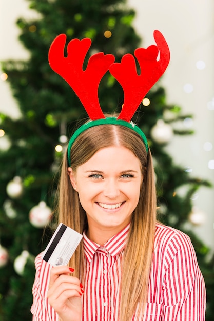 Free photo happy woman with plastic card in deer antlers headband near christmas tree