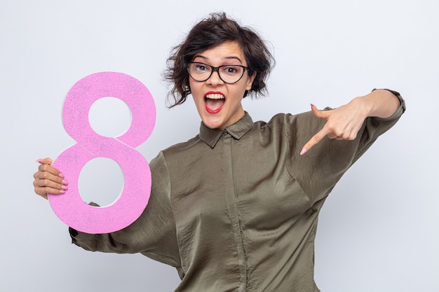 Free photo happy woman with short hair holding number eight made from cardboard pointing with index finger down smiling cheerfully celebrating international women's day march 8 standing over white background