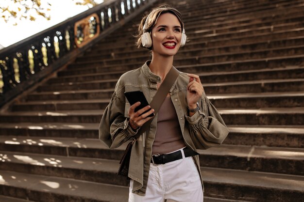 Happy woman with short hair and red lips in headphones smiles. woman in jacket and light pants holds phone outdoors.