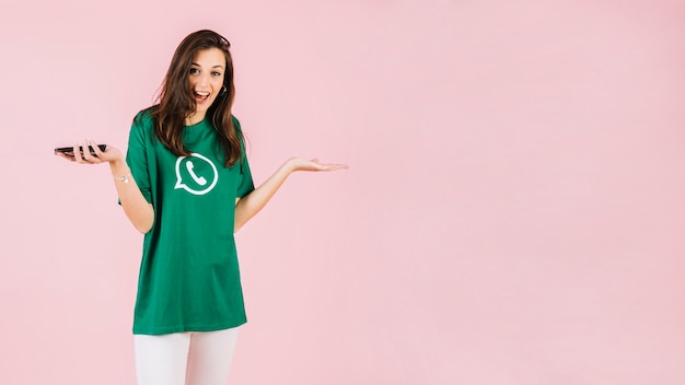 Free photo happy woman with smartphone shrugging on pink backdrop