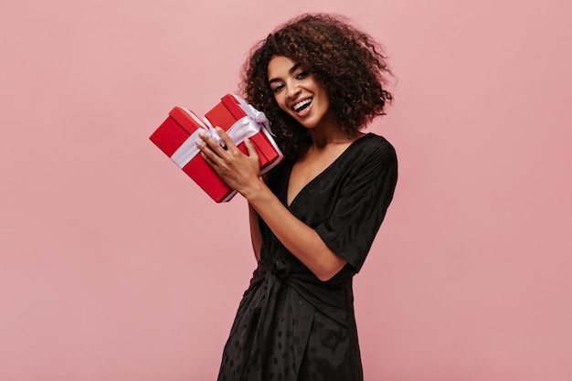 Happy wonderful mulatto woman with curly brunette hair in polk dot black dress smiling, looking into camera and holding two red gift boxes