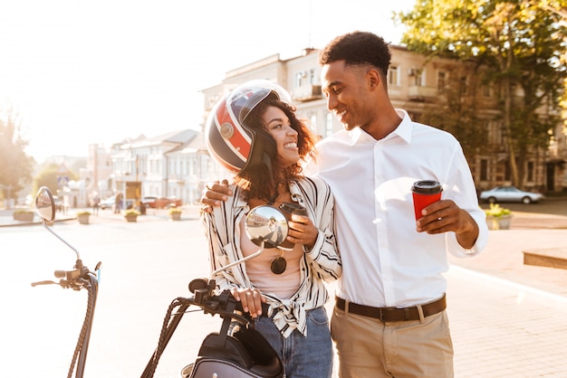 Happy young african couple drinking coffee while standing near the modern motorbike on the street and looking to each other